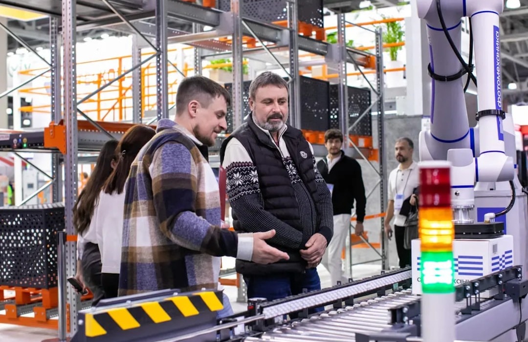 Attendees observe a robotic automation system at TRST Expo. Demonstration of advanced robotics for logistics and warehouse automation. A modern industrial exhibition featuring smart technology and innovation. | Men observe automated conveyor belt system in warehouse