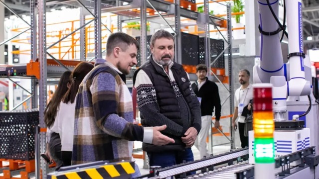 Attendees observe a robotic automation system at TRST Expo. Demonstration of advanced robotics for logistics and warehouse automation. A modern industrial exhibition featuring smart technology and innovation. | Men observe automated conveyor belt system in warehouse