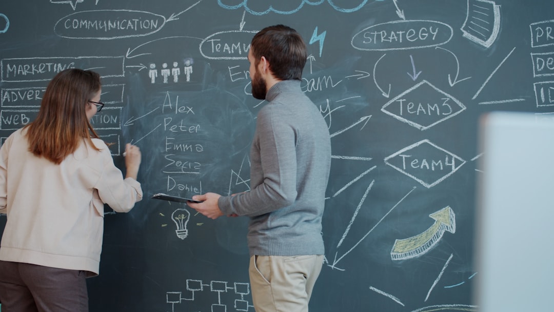 Woman employee is writing on blackboard talking to male coworker discussing business in open space office. Coworking, people and interior concept. | Two people planning on a chalkboard with diagrams.