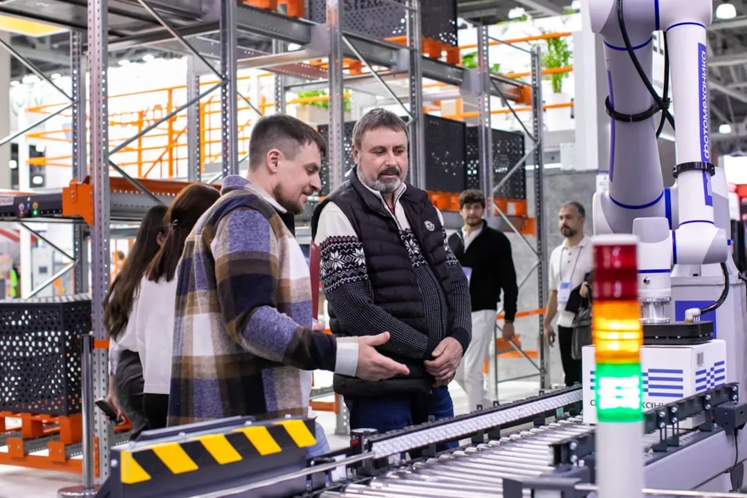 Attendees observe a robotic automation system at TRST Expo. Demonstration of advanced robotics for logistics and warehouse automation. A modern industrial exhibition featuring smart technology and innovation. | Men observe automated conveyor belt system in warehouse