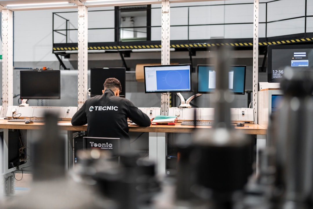Technician working on the design or analysis of mechanical components in front of several monitors at a computer station within an industrial plant. | A worker is concentrating at multiple computer screens.